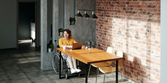 Woman in an orange cardigan in a wheelchair working from her laptop at a table looking out the window thinking and smiling.